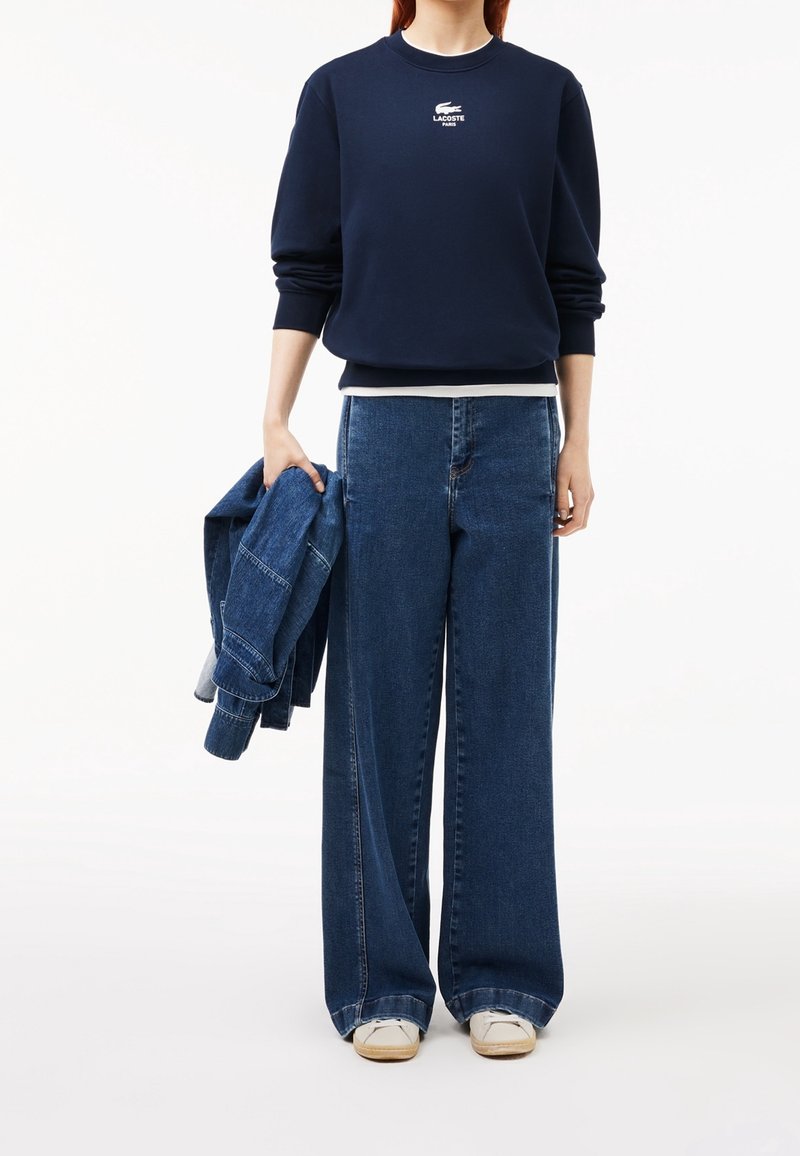 Personne portant un sweat-shirt bleu marine avec un logo blanc, un jean bleu à jambes larges, des baskets blanches, tenant une veste en jean pliée dans la main gauche.