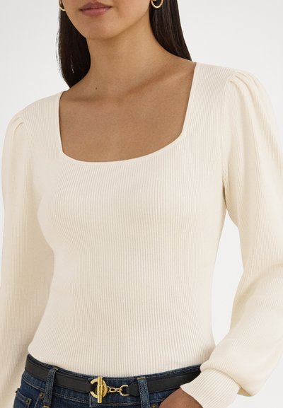 Haut crème côtelé à manches longues avec un décolleté carré et des manches bouffantes, associé à une ceinture sombre ornée d'un accessoire doré.