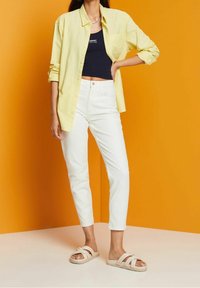 Camisa amarilla de botones sobre un top corto navy, pantalones ajustados blancos y sandalias beige en una modelo contra un fondo naranja.