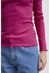 Tight långärmad topp i ribbad rosa tyg med en scalloped kant. Kombinerad med högmidjade ljusblå jeans.