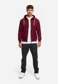 Burgundowa bluza z kapturem na zamek błyskawiczny z białymi sznurkami, z przednimi kieszeniami, nałożona na białą koszulkę, w zestawie z ciemnymi dżinsami i białymi sneakersami.