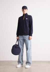 Hanorac cu fermoar de culoare navy, cu accente galbene, blugi drepți de culoare bleu deschis, adidași albi și o geantă rotundă de culoare navy. Design simplu, modern.