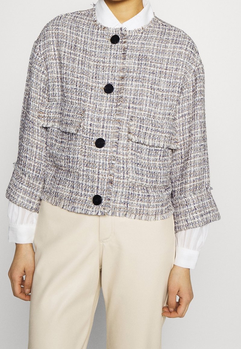 Veste en tweed tricotée au design court, présentant un motif texturé, quatre boutons noirs et deux poches avant. Col blanc en dessous visible.