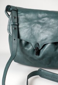 Borsa a spalla in pelle teal con chiusura a patta, dotata di clip nera, tracolla regolabile e superficie liscia. Include dettagli del logo sulla tracolla.