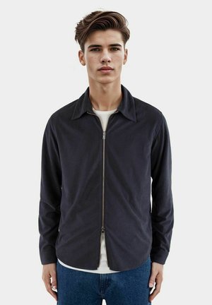 Marine blauwe zip-up jas met een kraagontwerp, lange mouwen, zijzakken en een gladde textuur, in laagjes over een wit shirt.