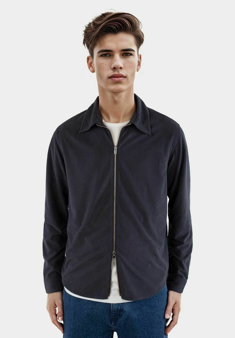 Giacca zip-up blu navy con design colletto, maniche lunghe, tasche laterali e una texture liscia, indossata sopra una camicia bianca.