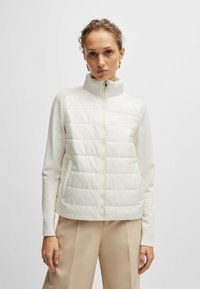 Gilet in piuma leggera di colore bianco sporco con colletto alto, parte anteriore trapuntata e maniche morbide. Presenta una zip completa e una silhouette aderente.