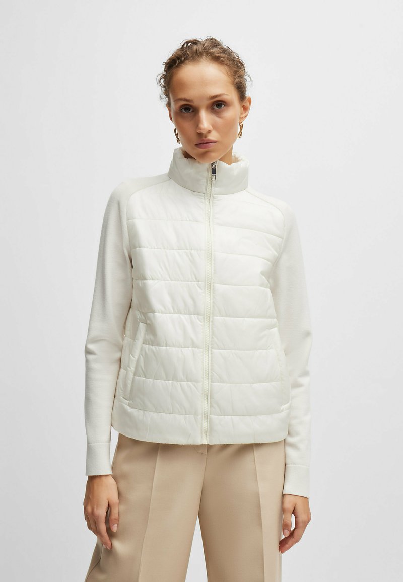Gilet in piuma leggera di colore bianco sporco con colletto alto, parte anteriore trapuntata e maniche morbide. Presenta una zip completa e una silhouette aderente.