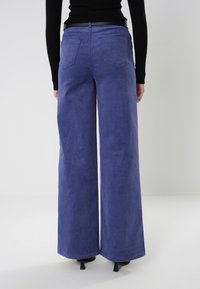 Persoon in hoogstaande, wijde blauwe corduroy broek, zwarte riem, zwart geribd langemouwen shirt en zwarte hakken, staande tegen een effen achtergrond.