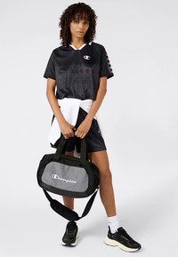 Zwart shirt van kort jersey met een ingewikkeld patroon, witte kraag, paarse accenten. Houdt een grijze en zwarte sporttas vast met het "Champion"-logo.