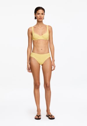 Femme debout sur fond blanc portant un bikini jaune texturé et des tongs noires, regardant droit devant avec une expression neutre.