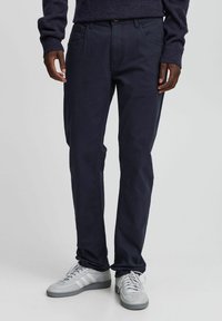 Navy chinos made of smooth fabric, featuring a straight fit, five-pocket design, and subtle stitching accents. Paired with light gray sneakers.