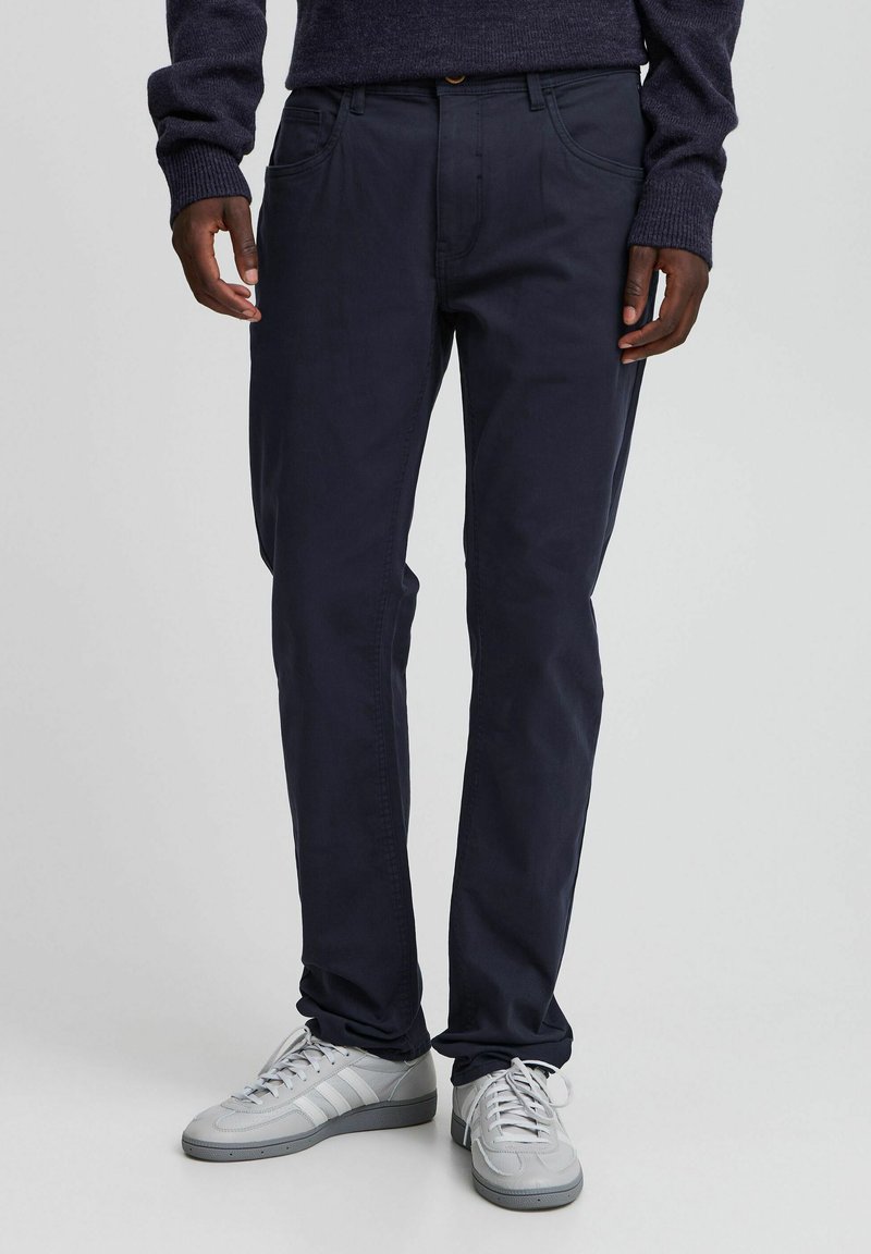 Navy chinos made of smooth fabric, featuring a straight fit, five-pocket design, and subtle stitching accents. Paired with light gray sneakers.