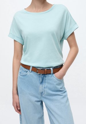 Femme portant un T-shirt bleu clair à manches courtes rentré dans un jean bleu clair avec une ceinture marron, une main dans la poche, sur un fond uni.