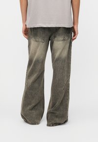 Άτομο που φοράει φαρδιά, ξεθωριασμένα μαύρα τζιν με φαρδιά μπάτζια και ανοιχτό γκρι μπλουζάκι, στέκεται μπροστά σε απλό λευκό φόντο.