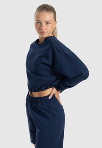 Felpa corta blu navy con maniche lunghe e orlo elasticizzato, abbinata a pantaloni shorts coordinati dotati di vita elasticizzata e tasche laterali.