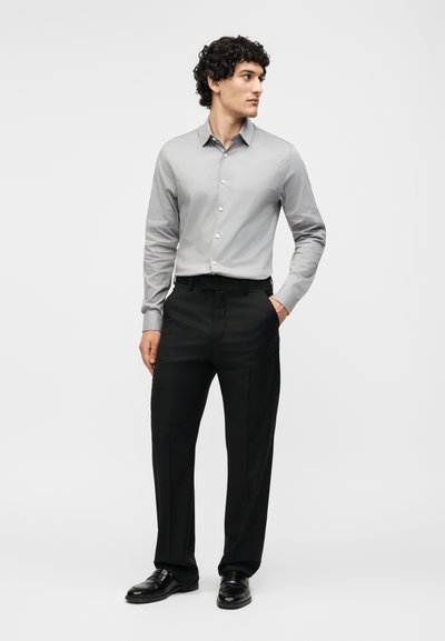 Άνδρας που στέκεται με το αριστερό χέρι στην τσέπη, φοράει ανοιχτό γκρι πουκάμισο με κουμπιά, μαύρο παντελόνι και μαύρα επίσημα παπούτσια, μπροστά σε απλό φόντο.