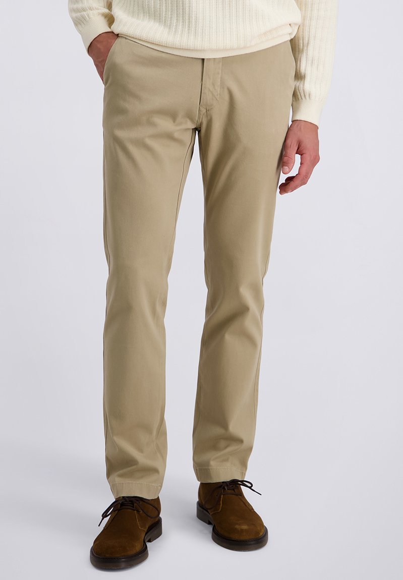 Mann in beigen Chinos, braunen Wildlederschuhen und einem cremefarbenen Rippstrickpullover, der mit einer Hand in der Tasche vor einem schlichten Hintergrund steht.