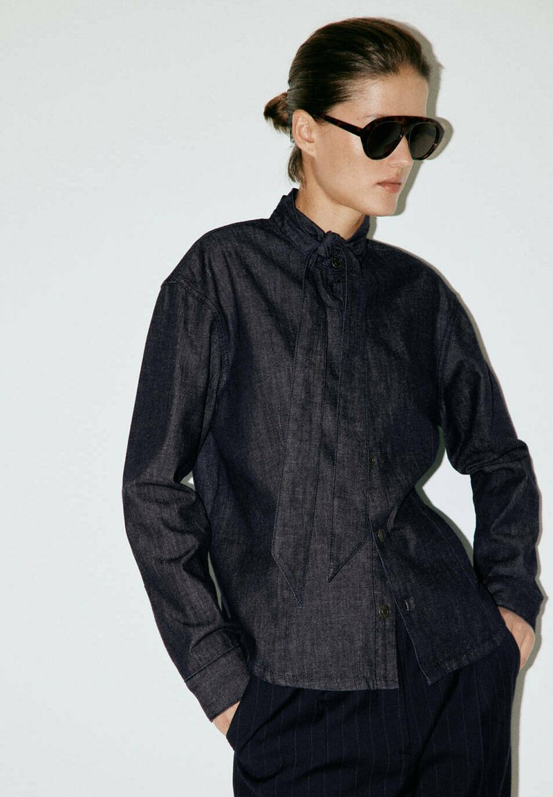 Femme portant une chemise en jean foncée boutonnée avec un détail de cravate au col, pantalon rayé foncé, grandes lunettes de soleil noires, mains dans les poches, regardant sur le côté.