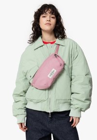 Giacca bomber verde chiaro, marsupio rosa con zip, materiale tessile. Pantaloni di jeans scuri, il modello indossa una maglietta rossa sotto la giacca.