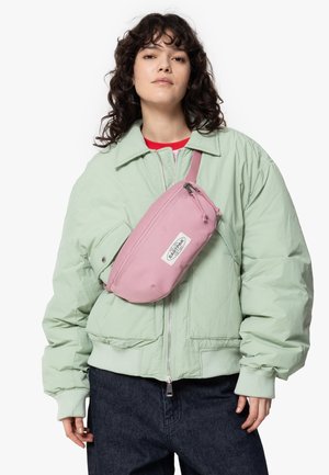 Light green bomber jacket, pink waist bag with zipper, textile material. Dark denim pants, model is wearing a red top under the jacket.