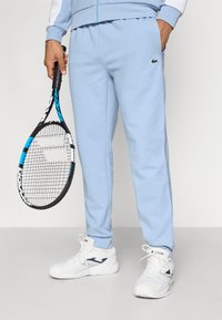 Ανοιχτό μπλε φόρμες με λωρίδες στο πλάι, ελαστική μέση και κεντημένο λογότυπο. Λευκά αθλητικά παπούτσια και ρακέτα τένις στο χέρι.