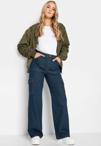 Oliivivihreä bombere takki, valkoinen t-paita ja leveälahkeiset tummansiniset cargo housut, joissa on kaksi sivutaskua ja erottuva ommelu. Valkoiset lenkkarit.