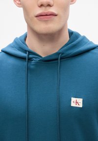 Mėlynas kapučinas su priekinėmis kišenėmis, raišteliais ir stačiakampiu Calvin Klein logotipu ant kairiojo krūtinės. Minkštas audinio paviršius.