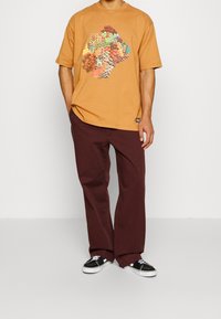 Orange grafisk t-shirt med abstrakt design, lös passform, parat med vinröda vida byxor och svarta sneakers med vita detaljer.