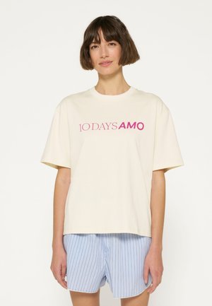 Crèmekleurig katoenen T-shirt met korte mouwen en een ronde halslijn, met "10 DAYS AMO" in het roze bedrukt. Gecombineerd met lichtblauwe gestreepte shorts.