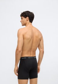 Homem com cabelo escuro curto, sem camisa, vestindo boxers pretos da Calvin Klein, de pé e olhando para a esquerda contra um fundo claro simples.