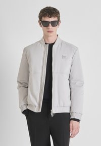 Jonge man met zwarte zonnebril, een lichtgrijze bomberjack met rits, zwart shirt en zwarte broek tegen een eenvoudige witte achtergrond.