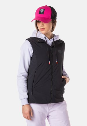 Gilet isolante nero con chiusura a zip, dotato di cappuccio e tasche. Abbinato a una felpa leggera di colore viola e a un berretto rosa e nero.