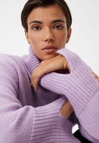 Lila Strickpullover mit hohem Kragen und offenem Ärmeldesign, der ein strukturiertes Muster und ein weiches, warmes Aussehen präsentiert.