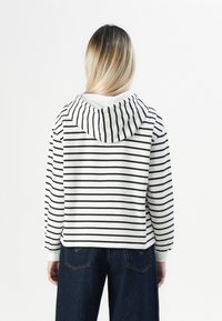 Personne avec de longs cheveux blonds portant un sweatshirt blanc avec des rayures horizontales noires et un jean bleu foncé, tourné le dos sur un fond blanc.