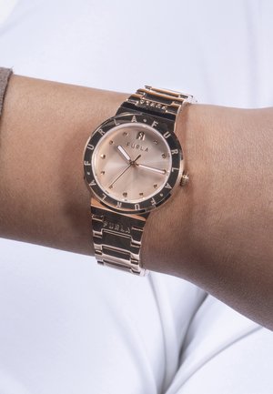 Auksinės spalvos metalinis laikrodis su rožinės aukso ciferblatu, turintis poliruotą paviršių, taškinius žymeklius ir išgraviruotą "Furla" detalę ant rėmelio ir diržo.