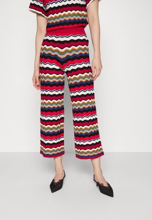 Vrouw draagt kleurrijke zigzag-gepatroonneerde broek en bijpassende top, gecombineerd met zwarte pumps met spitse neus en kanten detail.