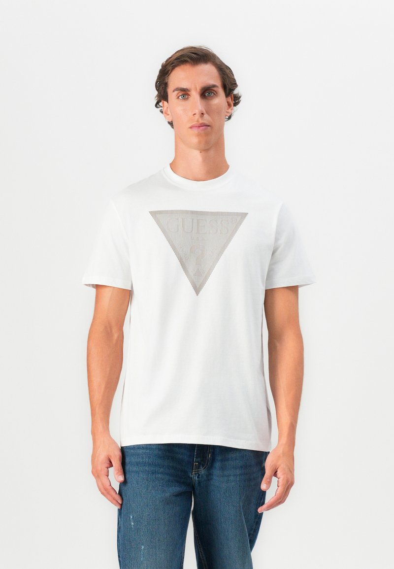 Jeune homme portant un t-shirt blanc avec un graphique en triangle gris et un jean bleu, se tenant devant un fond blanc uni.