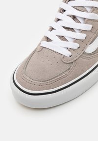 Beige semsket sko med hvite snørebånd, med en hvit sidestripe, rund tå, svart kant, og teksturert gummisåle.