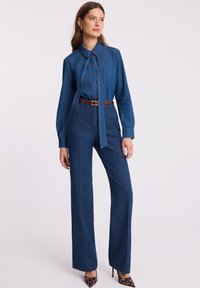 Kobieta ubrana w niebieską dżinsową bluzkę z wiązaniem pod szyją oraz dżinsy o wysokim stanie i szerokich nogawkach, z brązowym paskiem oraz obcasami w leopardzi wzór, stojąca na białym tle.