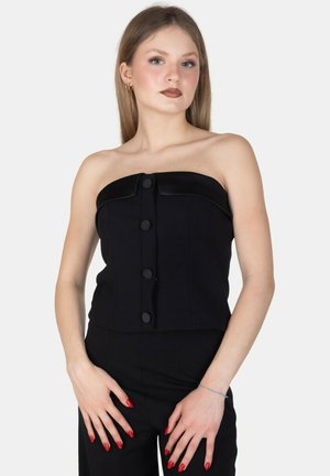 Giovane donna che indossa un top nero senza spalline con bottoni e pantaloni neri coordinati, posa con smalto rosso su sfondo chiaro.
