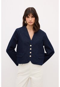 Chaqueta de mezclilla azul marino con escote en V, mangas largas, dos bolsillos frontales y cinco botones de perlas en la parte delantera. Textura suave y ajuste entallado.