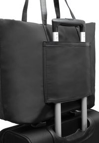 Sac fourre-tout noir fixé à la poignée d'une valise noire à roulettes avec poignée télescopique déployée sur fond blanc.