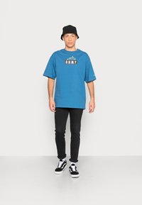 T-shirt en coton bleu à manches courtes avec un logo graphique "GRMY", associé à un jean skinny noir et des baskets noires avec des accents blancs.