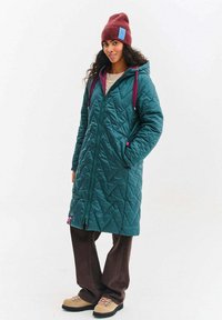 Tyrkysově modrá prošívaná dlouhá bunda s kapucí, zapínáním na zip, vínovými doplňky a růžovým logem. Kombinována s hnědými kalhotami a světlými botami.
