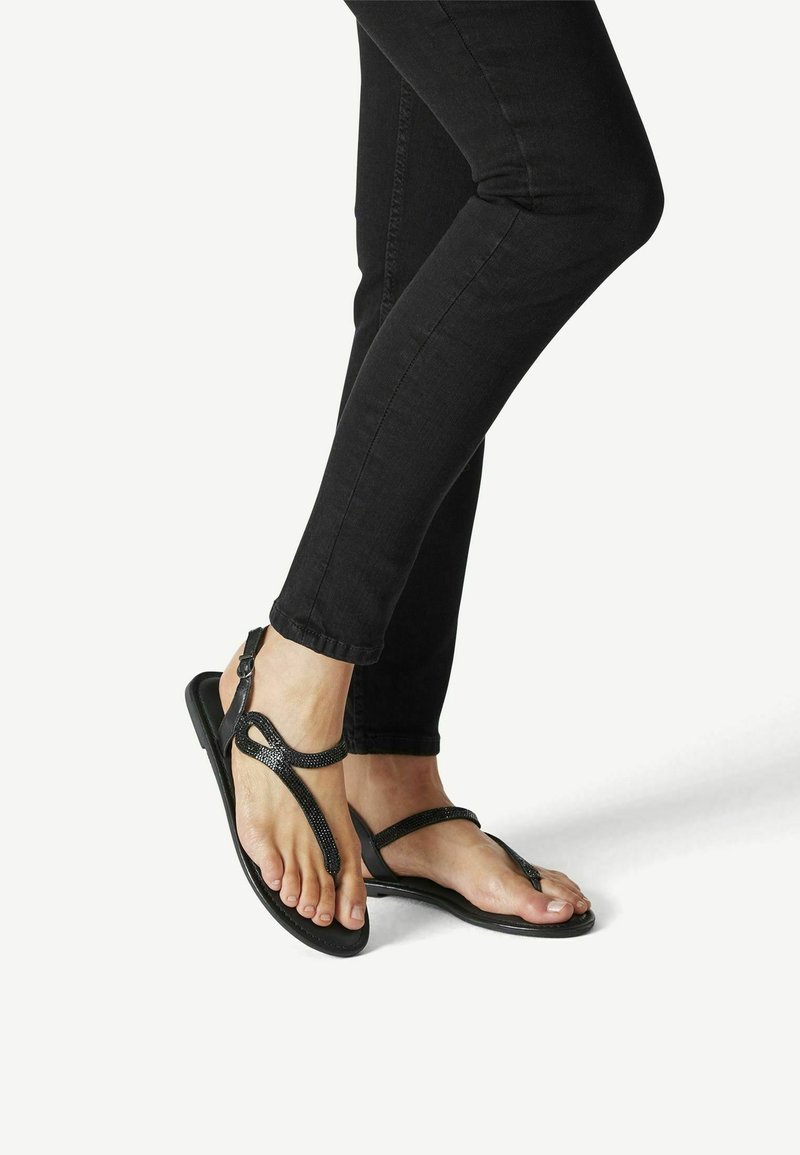 Sandalias negras con suela plana, con un diseño de tiras texturizadas y hebilla en el tobillo. Llevadas con jeans ajustados negros.