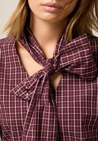 Bordeaux geruite blouse met een geknoopte halsafwerking, voorzien van een kleine sleutelgaatje uitsnijding en versierde knopen op de kraag voor extra accent.