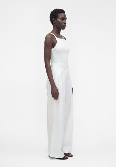 Femme debout en haut blanc sans manches et pantalon large taille haute avec des sandales noires sur un fond blanc uni.