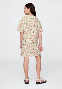 Mergina stovinti nugarą pasukusi, dėvinti kreminės spalvos trumpomis gėlėmis margintą suknelę ir baltas basutes ant paprasto fono.