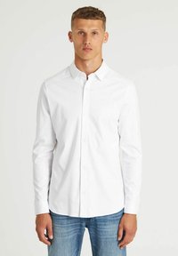 Weißes langärmliges Hemd mit Button-Down-Kragen, aus Baumwolle, mit einem dezenten Logo auf der Brust und geradem Saum. Getragen mit blauen Jeans.